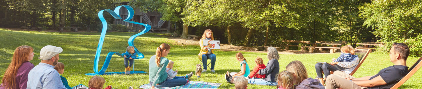 Auf einer grünen Wiese im Park sitzt eine Gruppe junger Eltern mit Kinder. Vor ihnen sitzt eine Lehrerin und erzählt eine Geschichte aus einem Buch. Ein kleiner Junge klettert auf dem menschengrossen blauen Stiftungslogo aus Metall herum, das links neben der Gruppe auf der Wiese steht.